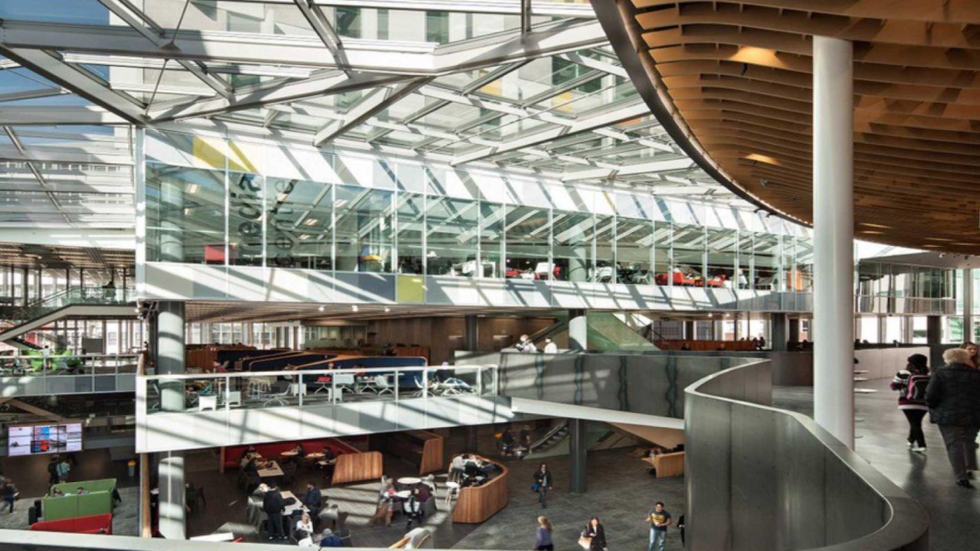 A modern, open-concept building with large glass windows, multiple floors, and people walking or sitting in common areas. The space features bright natural light and contemporary architectural elements.