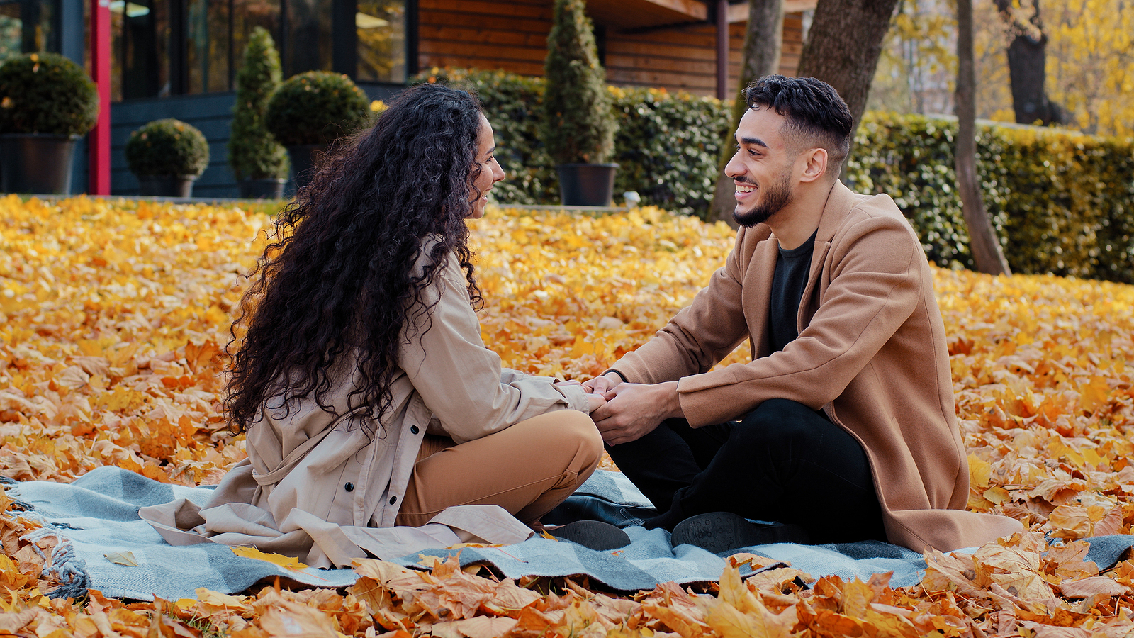 Happy Hispanic couple on a date in an autumn park. They are young, holding hands, laughing, and smiling while sitting cross-legged on a plaid blanket amidst colorful leaves outdoors. The bearded guy is emotionally talking with his curly-haired girlfriend.