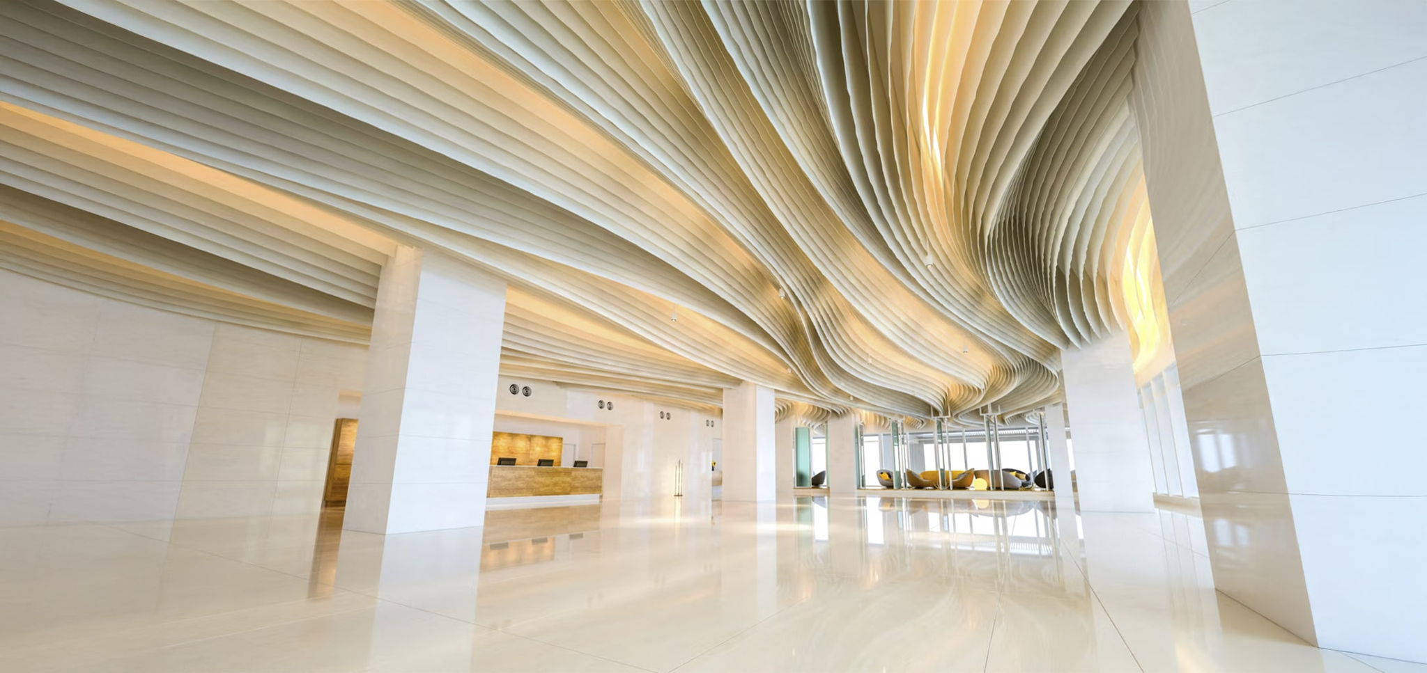 A modern, spacious lobby with glossy floors and dramatic, wave-like ceiling panels. The reception desk is on the left, and natural light fills the area through large windows at the far end.