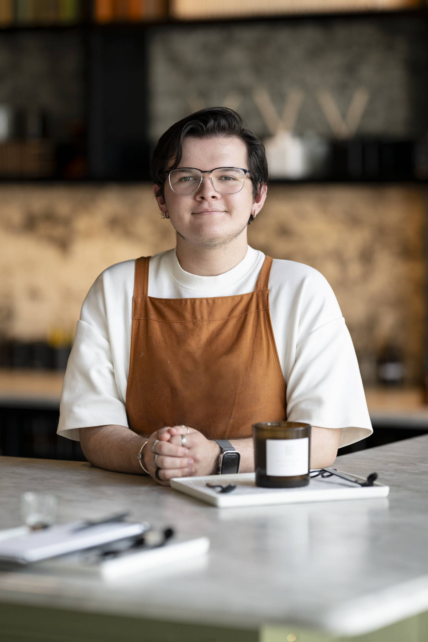 man with glasses in orange apron standing at counter in front of candle bar