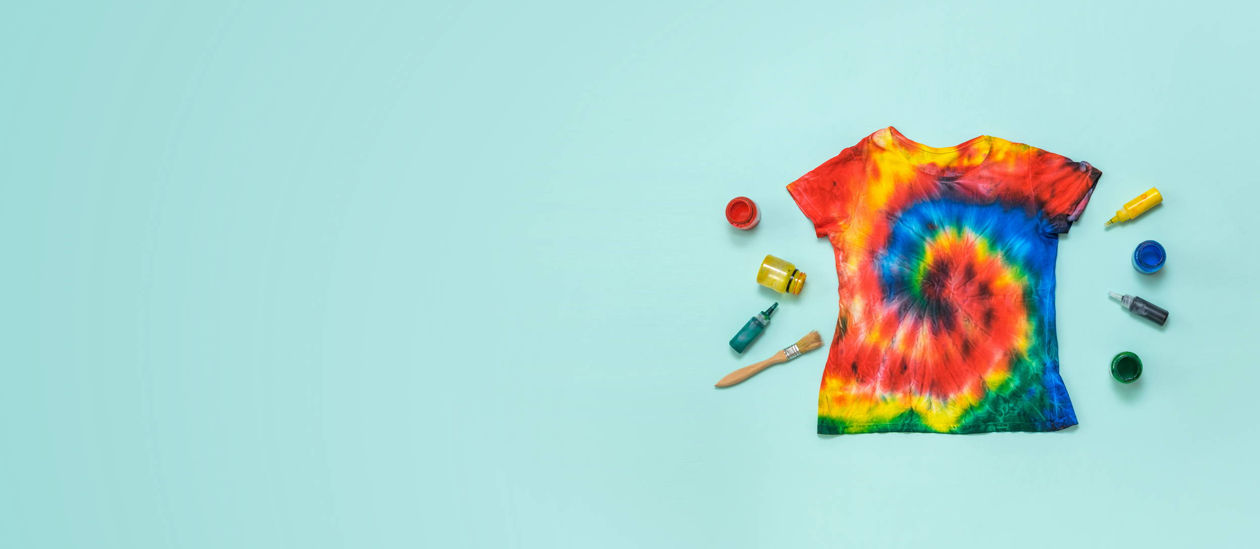 A tie dyed t-shirt surrounded by colorful ink dropper bottles and a paint brush for Confetti's Virtual Tie Dye Class