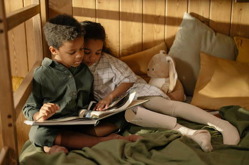 2 children, a girl and a boy, cozying up in bed while reading a book - Photo by cottonbro from Pexels