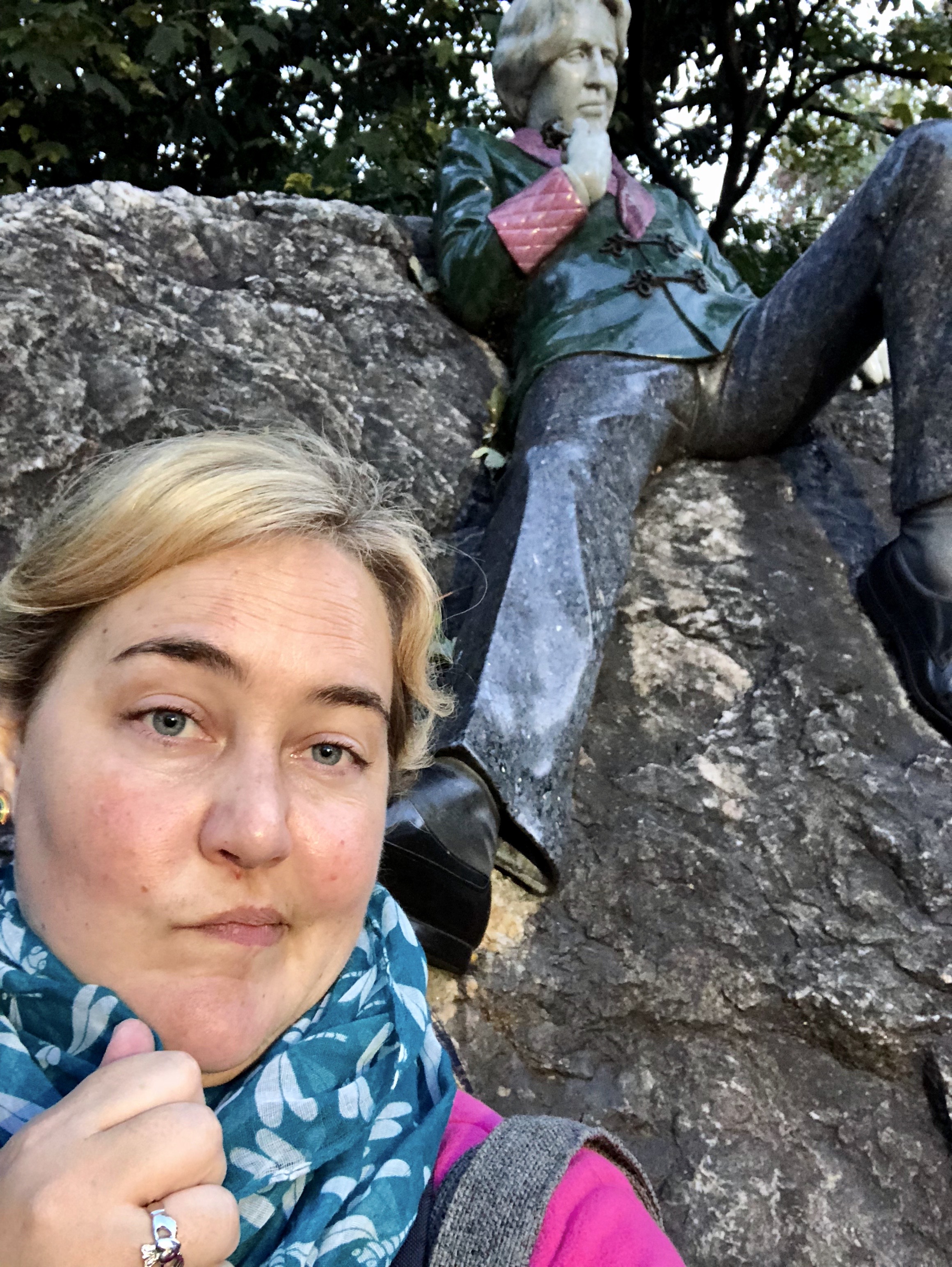 Jennie posing with a smirk in front of the Oscar Wilde statue sitting on top of a rock.