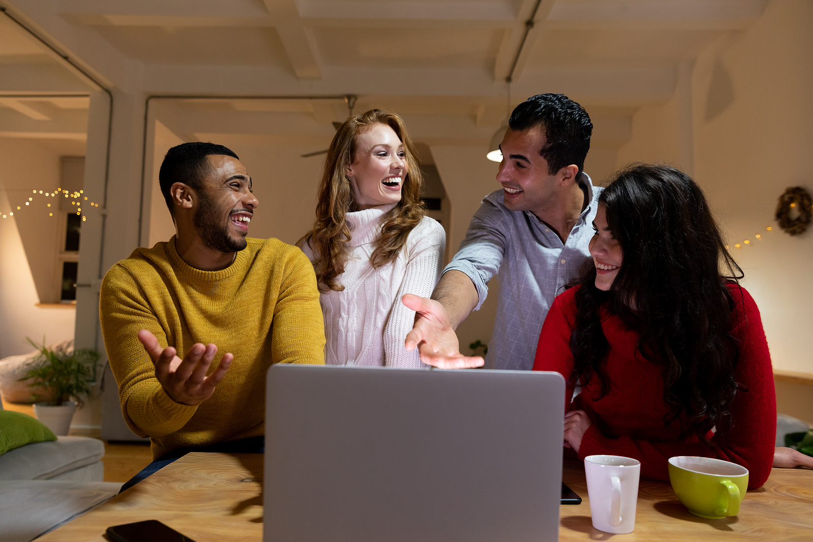 A group of four young multi-ethnic friends looking at a laptop computer together, talking and laughing in the sitting room of an apartment in the evening.