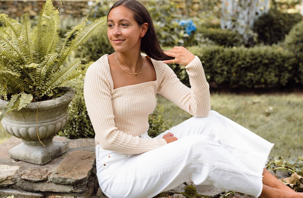 Cait Khosla, founder of Botany Box, lounges outside amongst plants in a white top and white jeans
