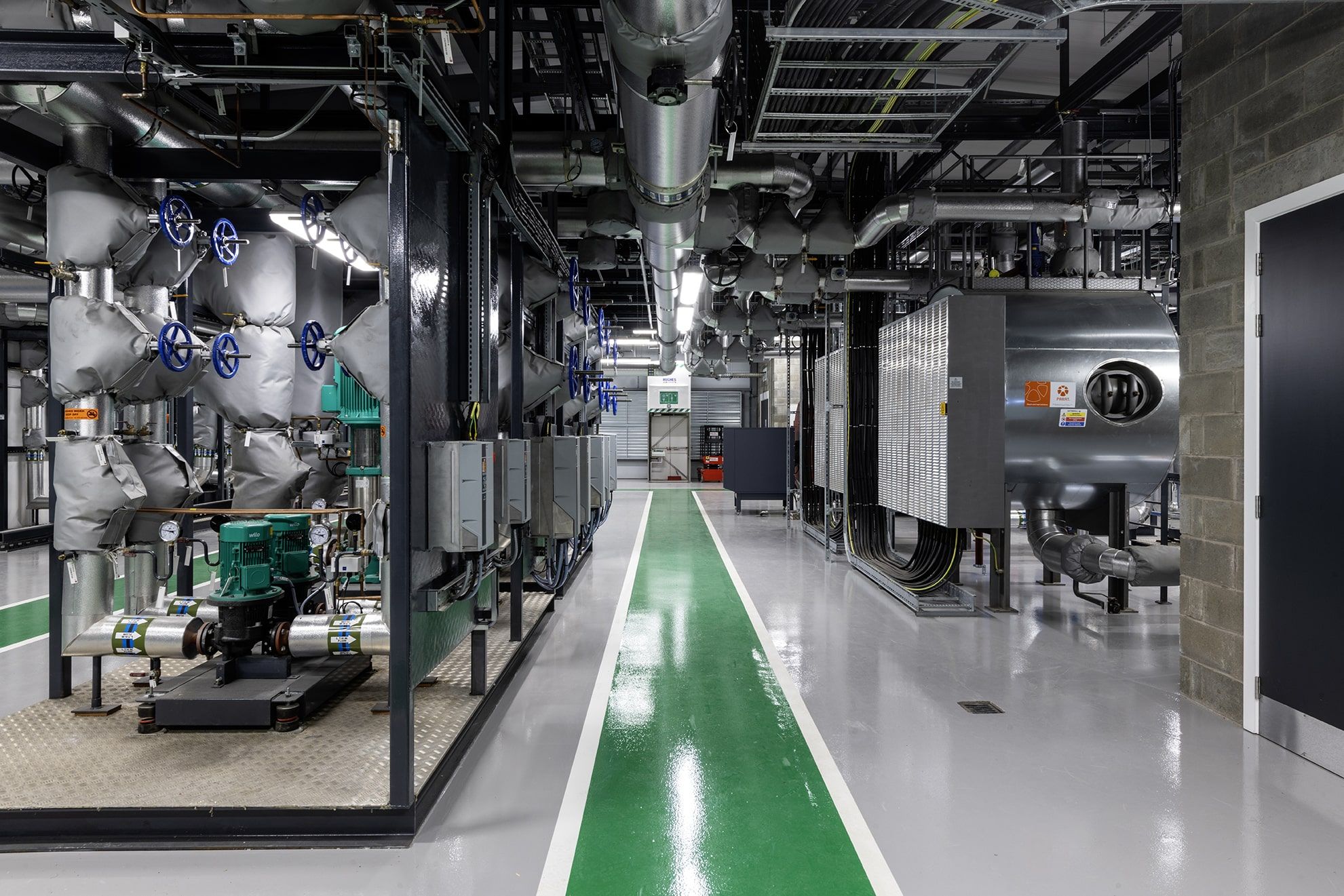 A modern industrial facility interior with machinery, pipes, and control panels. The floor has green and white painted paths, and the area is brightly lit with a clean, organized appearance.