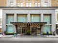 Die moderne Steinfassade des Hotels 'ROW NYC' mit großen Glasfenstern, beleuchtetem Vordach, Drehtüren und bepflanzten Kübeln.