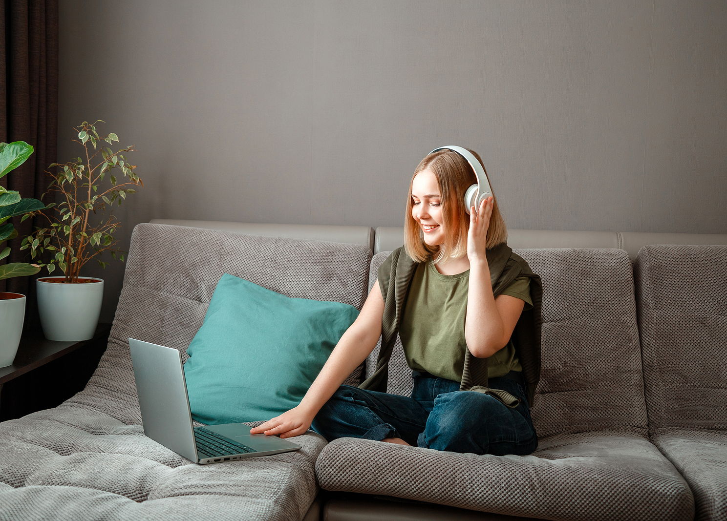 A happy young woman is sitting on sofa with laptop in headphones talking to friends online.