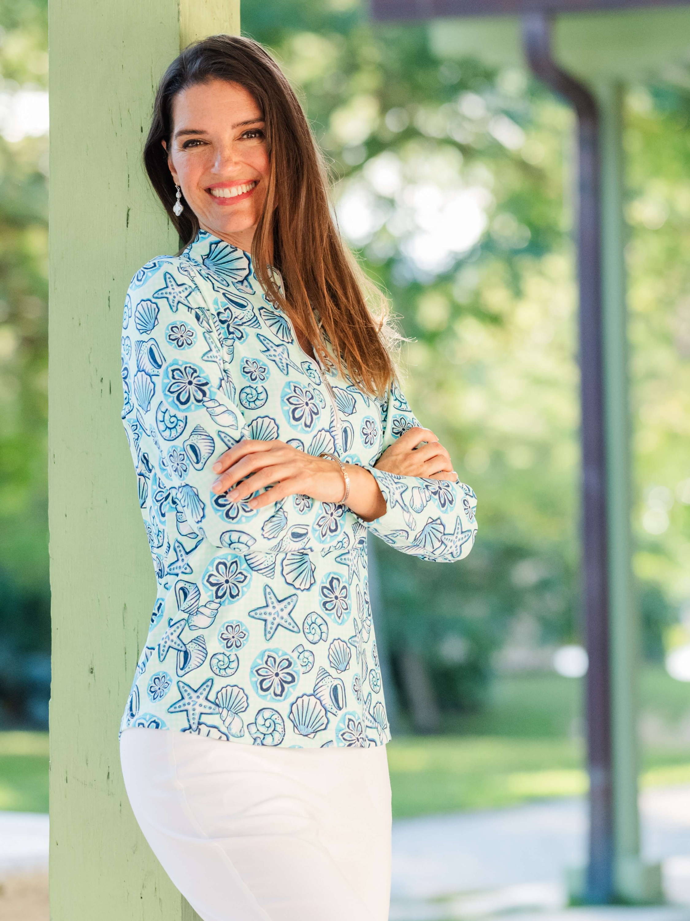 A woman wearing a green and blue printed sun shirt and white pants crosses her arms in front of her chest and leans against a wooden post in a park pavillion