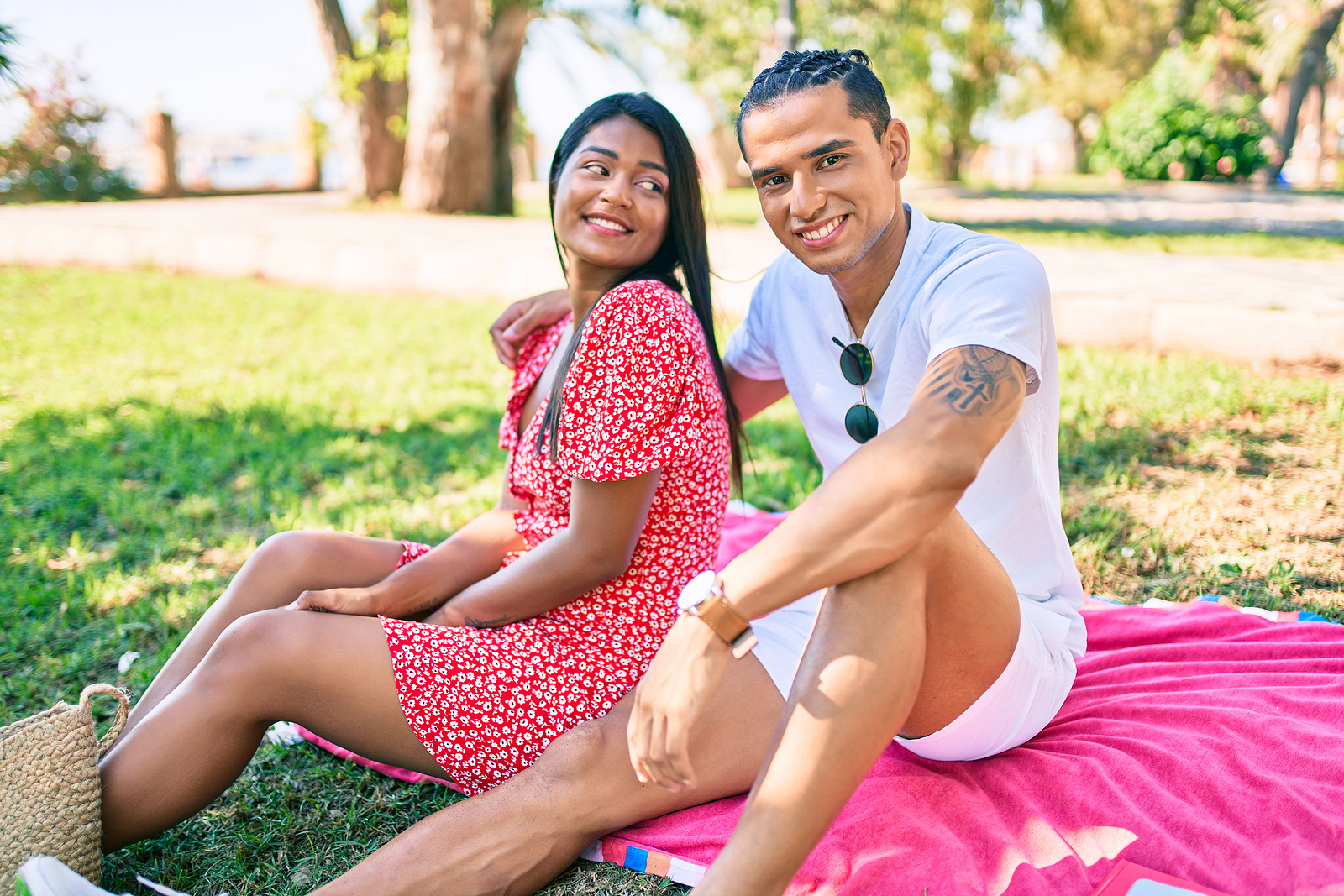 A young man with an arm around a smiling young woman. They're sitting on a blanket outdoors.