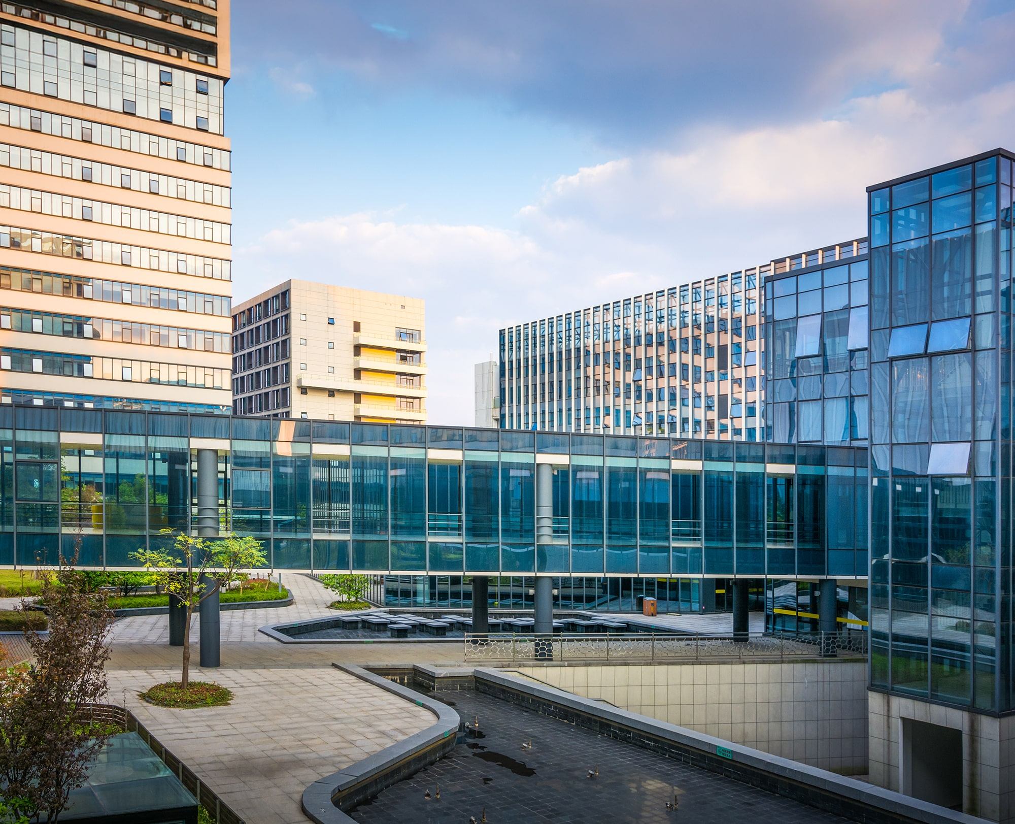 Modern office buildings with glass facades surround a courtyard featuring landscaped greenery, pathways, and a dry fountain under a partly cloudy sky.