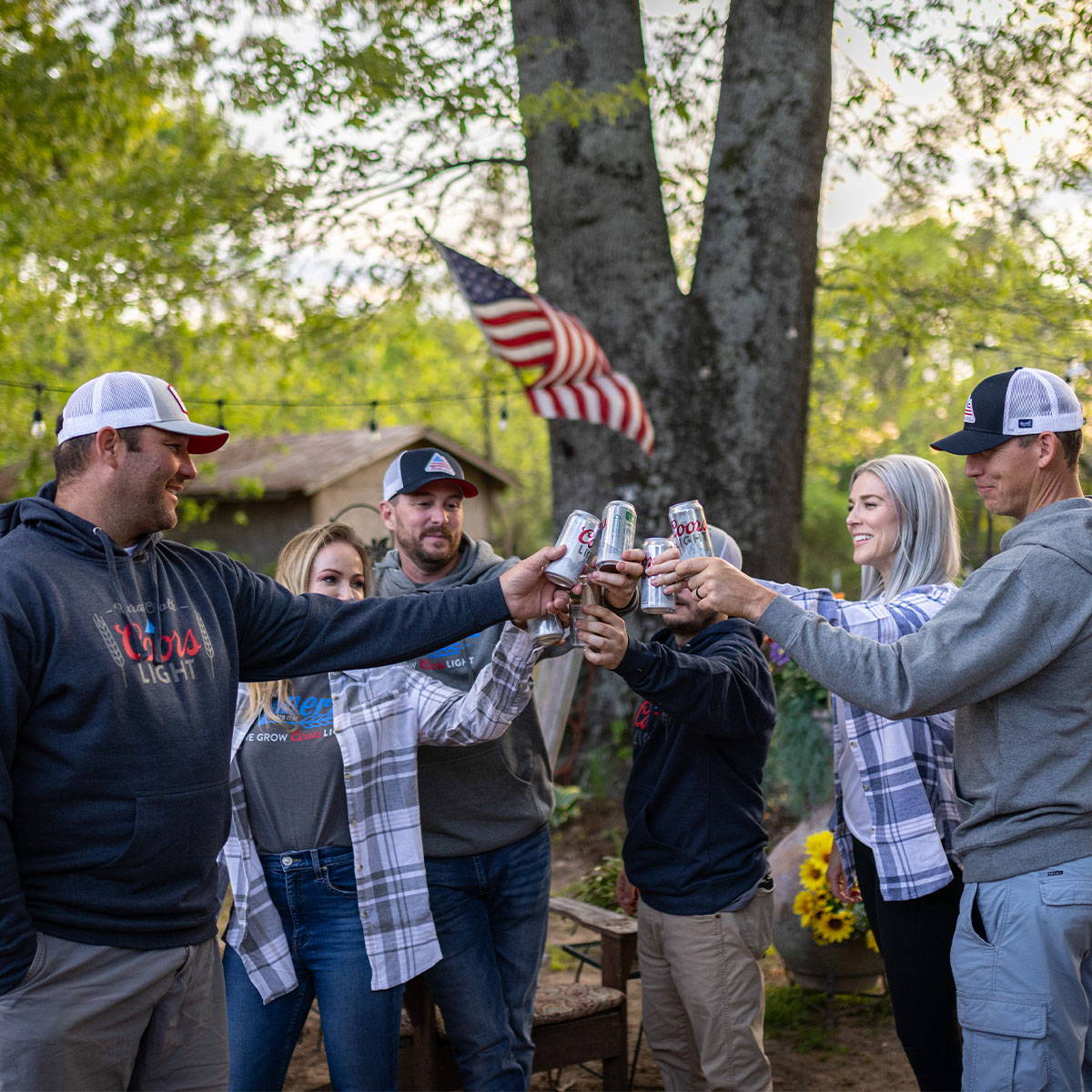 A group of people smiling and raising beers together wearing Coors Light Clothing. This represents the Rural Cloth and Coors Light Collection