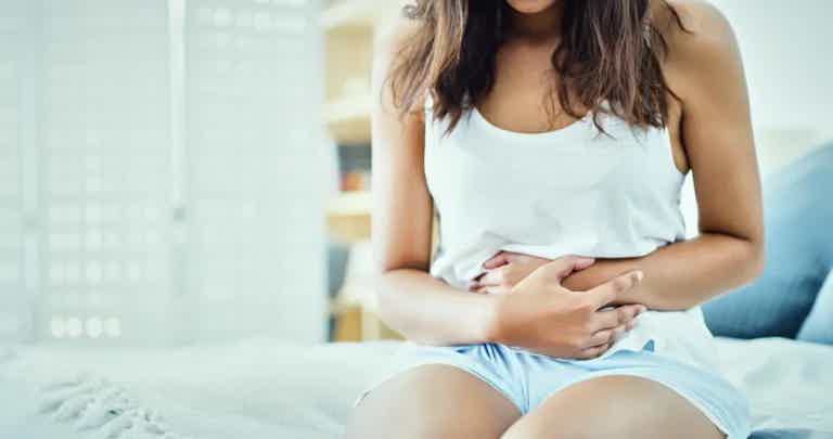 Woman sitting on a bed with hands on stomach, wearing a white tank top and light shorts, appearing to feel abdominal discomfo