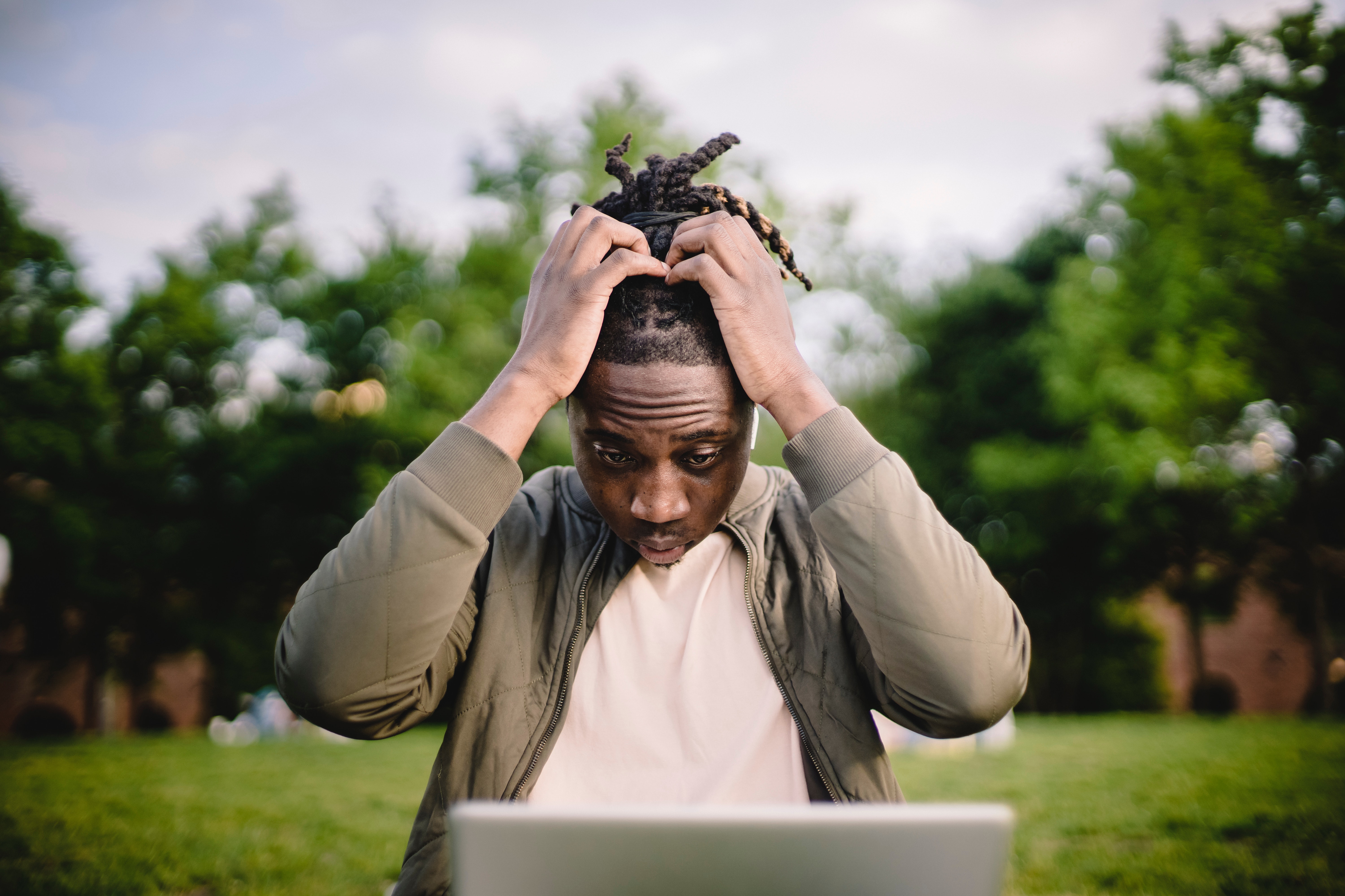 Image of young black man looking at his laptop in a park with his hands in his head looking stressed and breathing out.