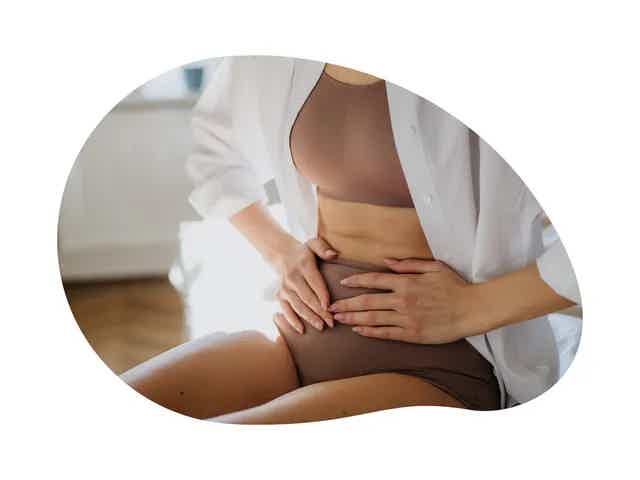 Person in white open shirt touching abdomen, wearing brown underwear, seated with hands on stomach.