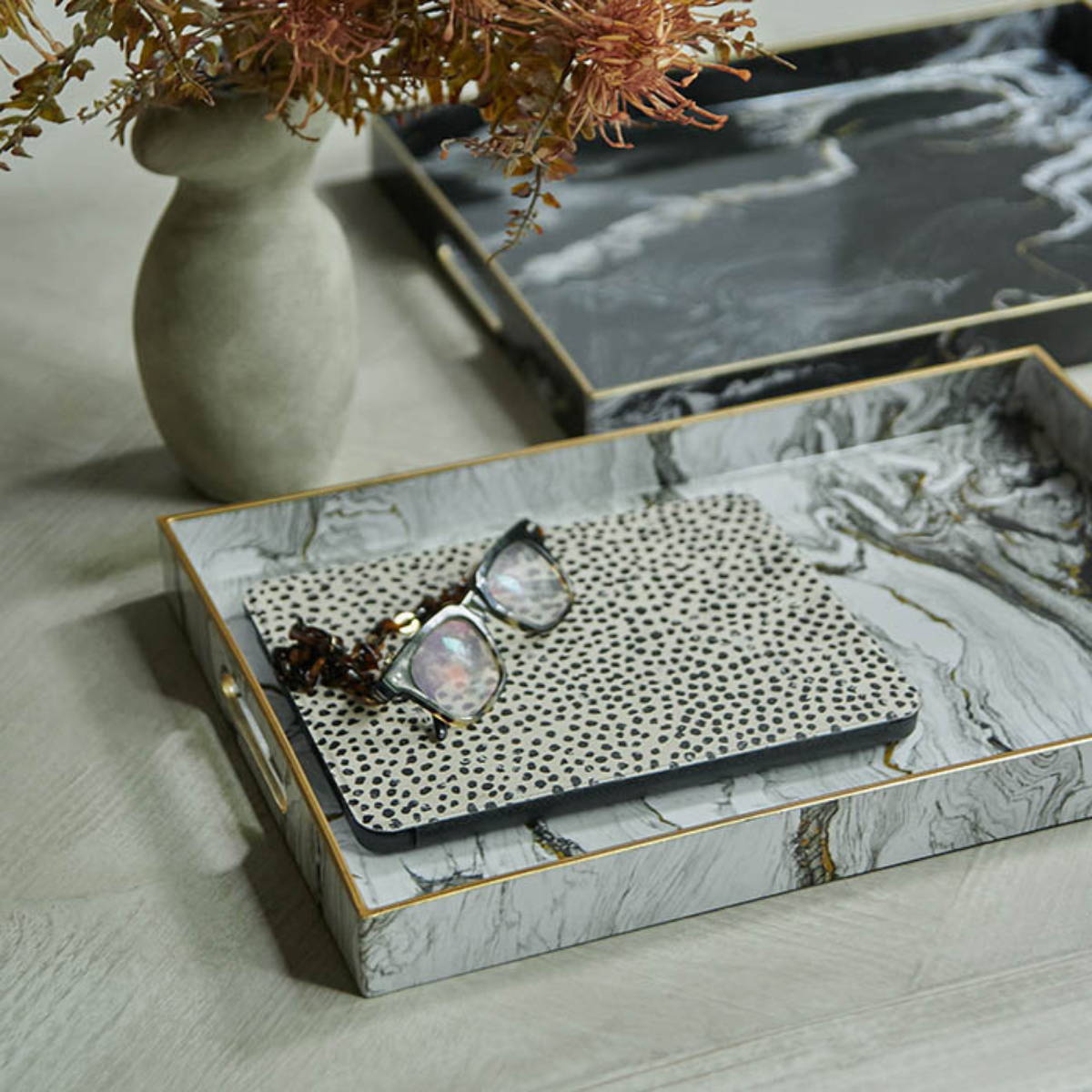two trays with a marble pattern holding a pair of reading glasses with a dotty notebook and a shapely vase with artificial flowers