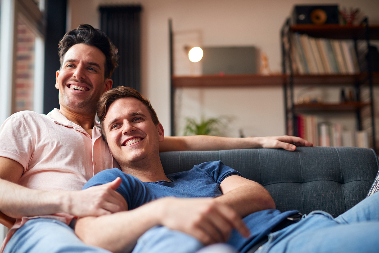 An attractive gay couple is cuddling on the couch together smiling watching tv.