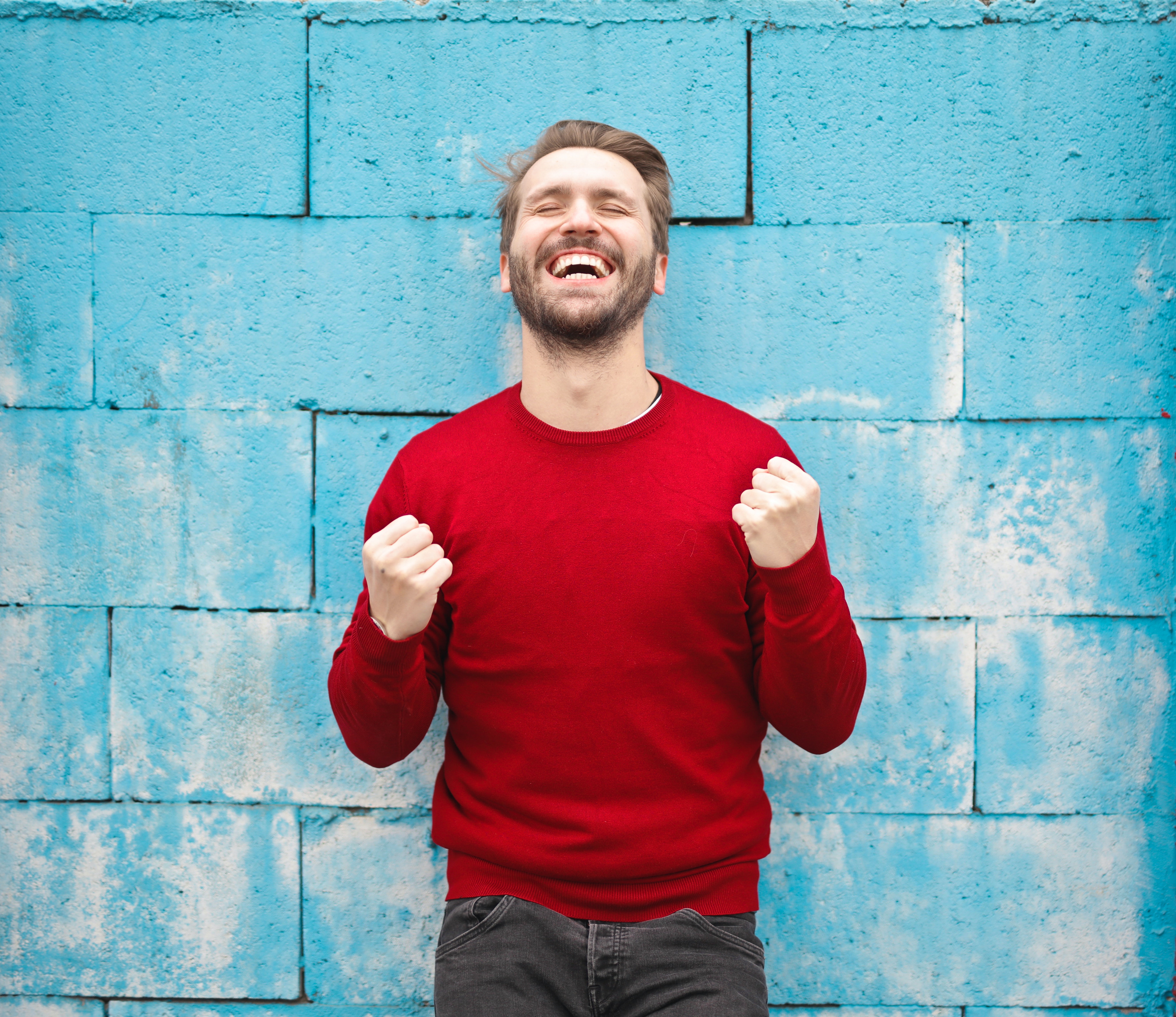 Image of a white man with a beard, wearing a bright red sweater on a light blue brick background, smiling with his eyes closed and his hands clenched near his chest.