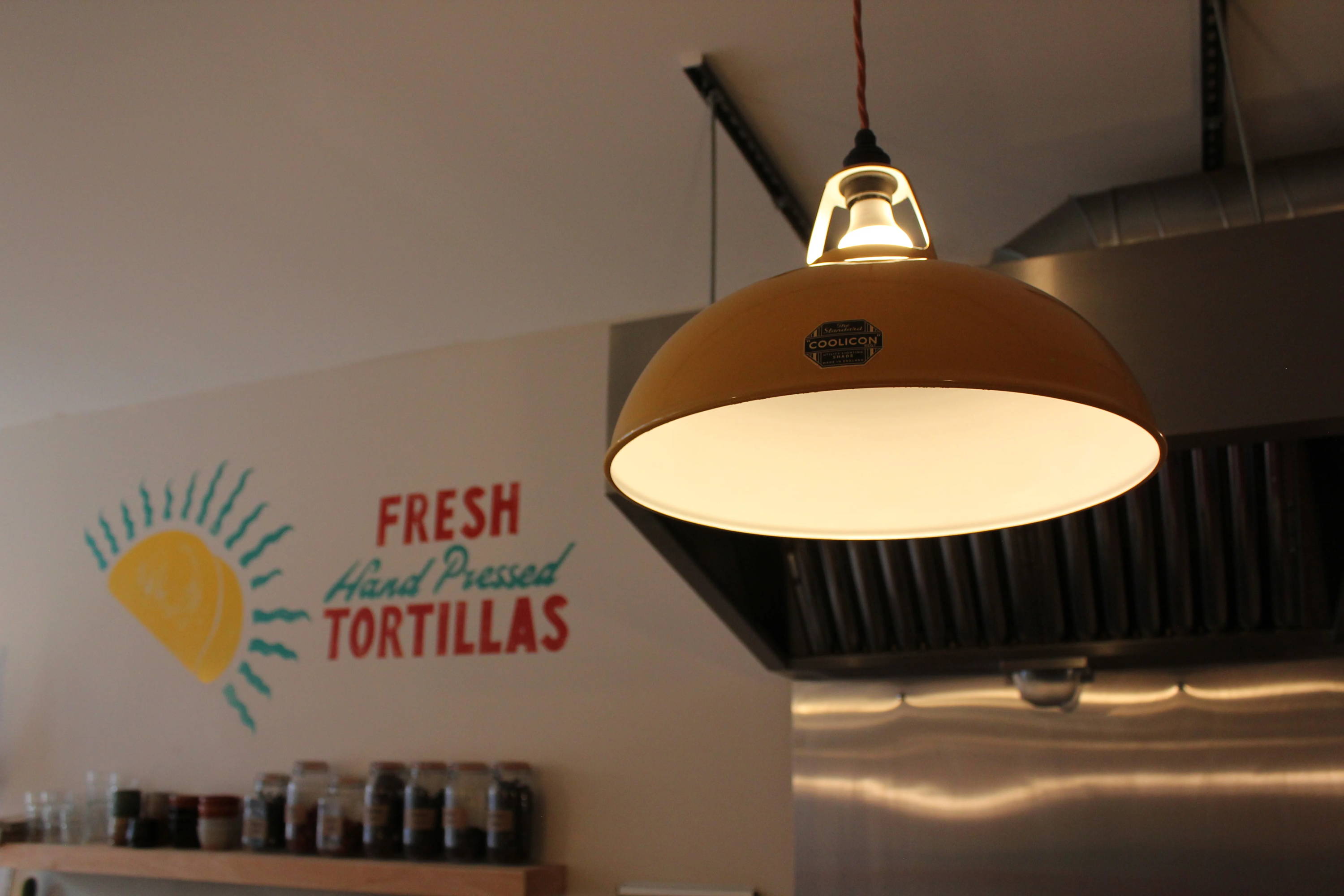 A close-up of a large Yellow Lampshade with an LED bulb and Black Lampholder with red cable. The Coolicon badge is cleary in focus. In the background is a hand-painted sign on the wall with an industrial steel grill.