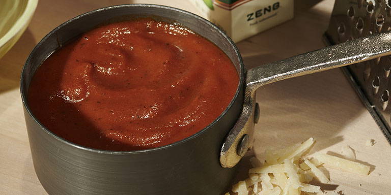 An anodized aluminum pan full of rich tomato sauce sitting on a wood surface with a grater, grated cheese and a carton of ZENB Gourmet Sauce in the background.