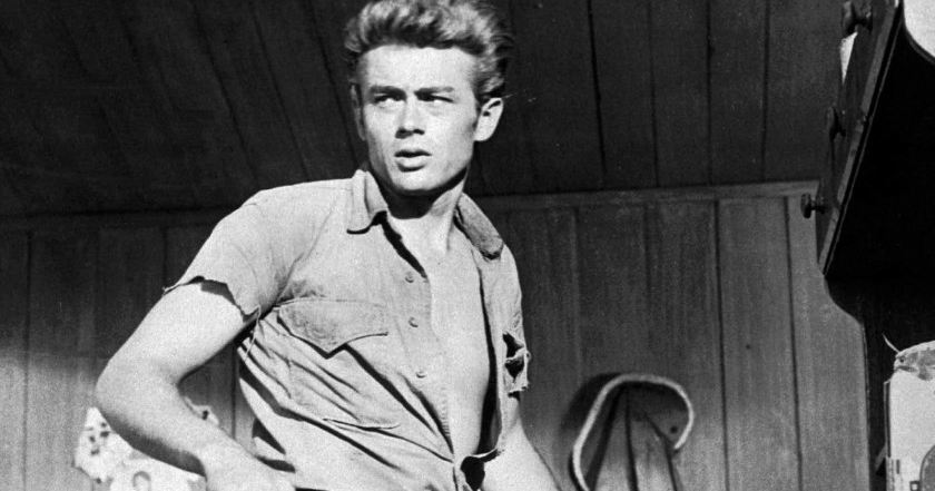 Black and white image of James Dean with his shirt unbuttoned as he looks to his side.
