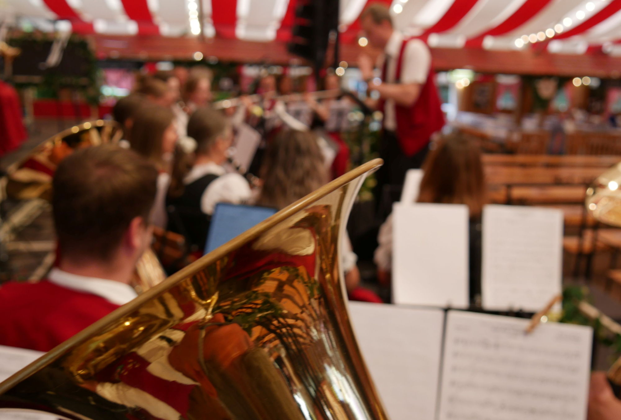 Nahaufnahme eines Tenorhorns vor einem Notenständer. Im Hintergrund ist eine Blaskapelle mit Dirigent zu sehen, die in einem Festzelt spielt. 