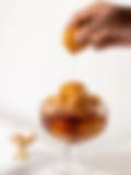 A hand holding a syrup-dripping gulab jamun over a bowl of golden gulab jamuns, with a small brass lamp in the background.