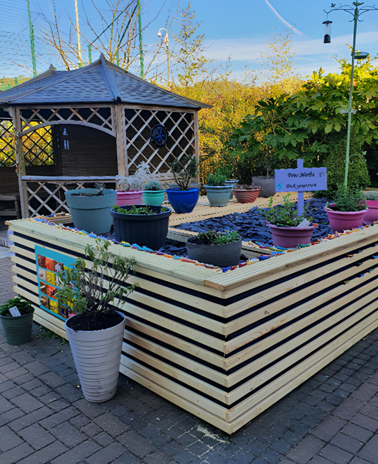 A raised wooden planter filled with soil and various potted plants stands on a paved area, with a wooden gazebo and greenery in the background. A sign reads Grow Herbs with a list of herb names.