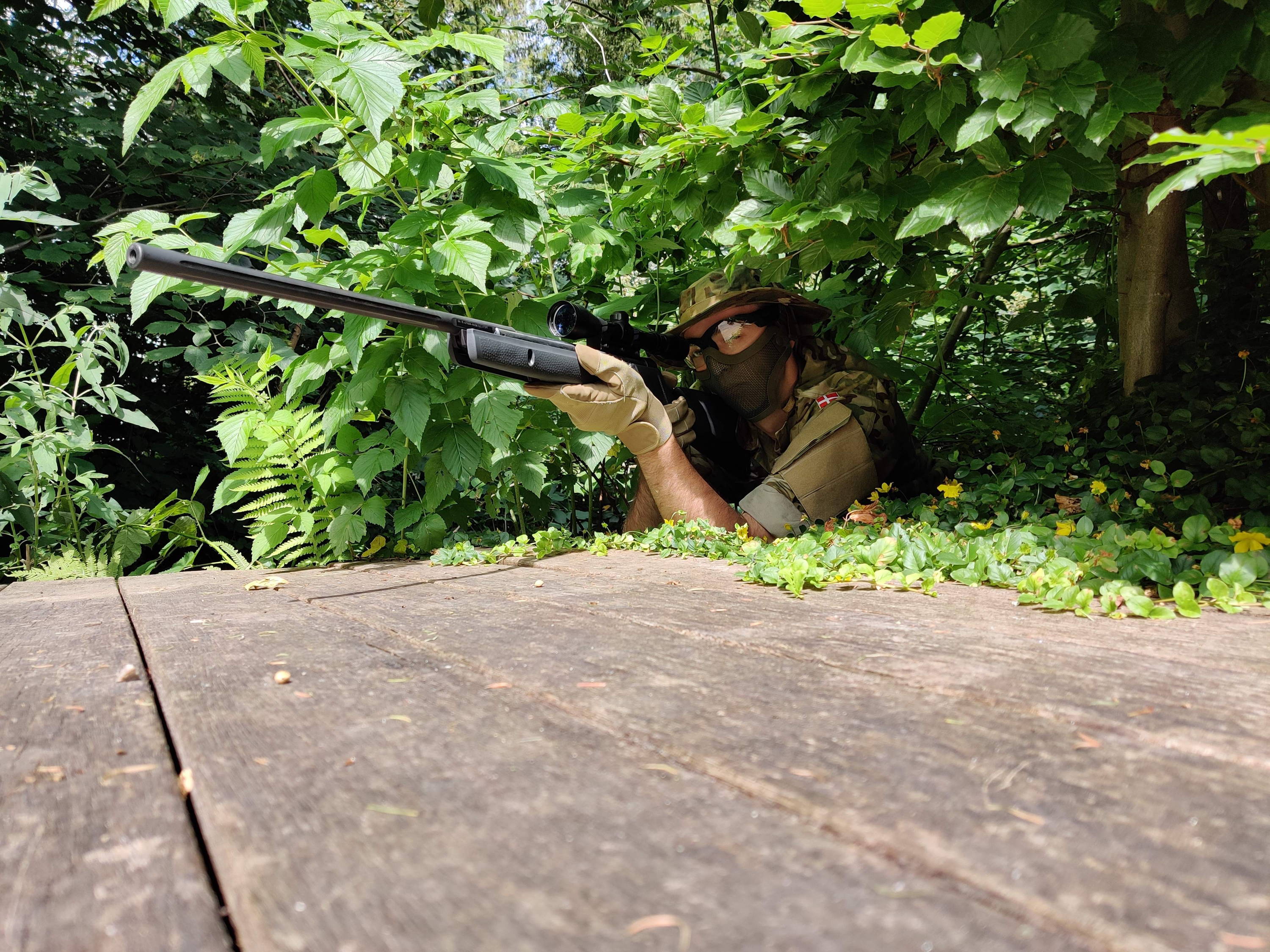Billede af en mand med en sniper som ligger i en busk med camouflagetøj og mercury kamera skydebriller