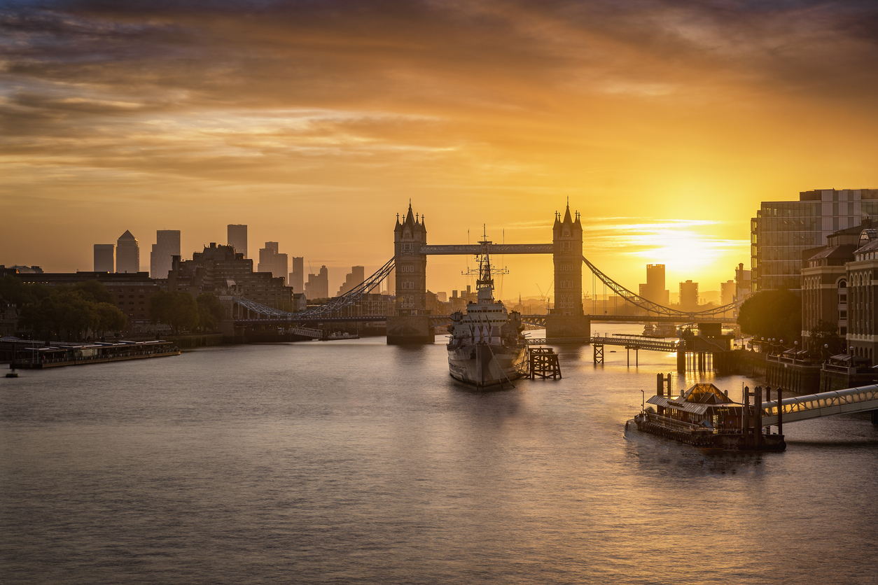 Tower Bridge over the River Thames in London at sunrise, with a moored ship in the foreground and the sun casting a golden glow across the sky and water.