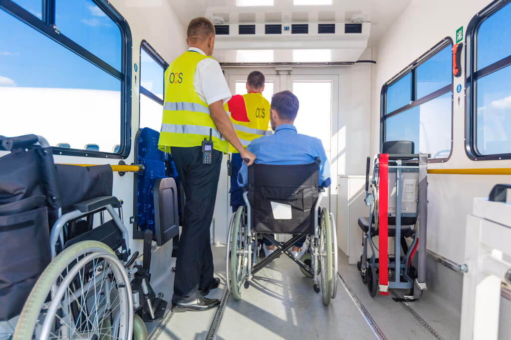 A person in a wheelchair is assisted onto a vehicle by staff wearing reflective OCS vests.