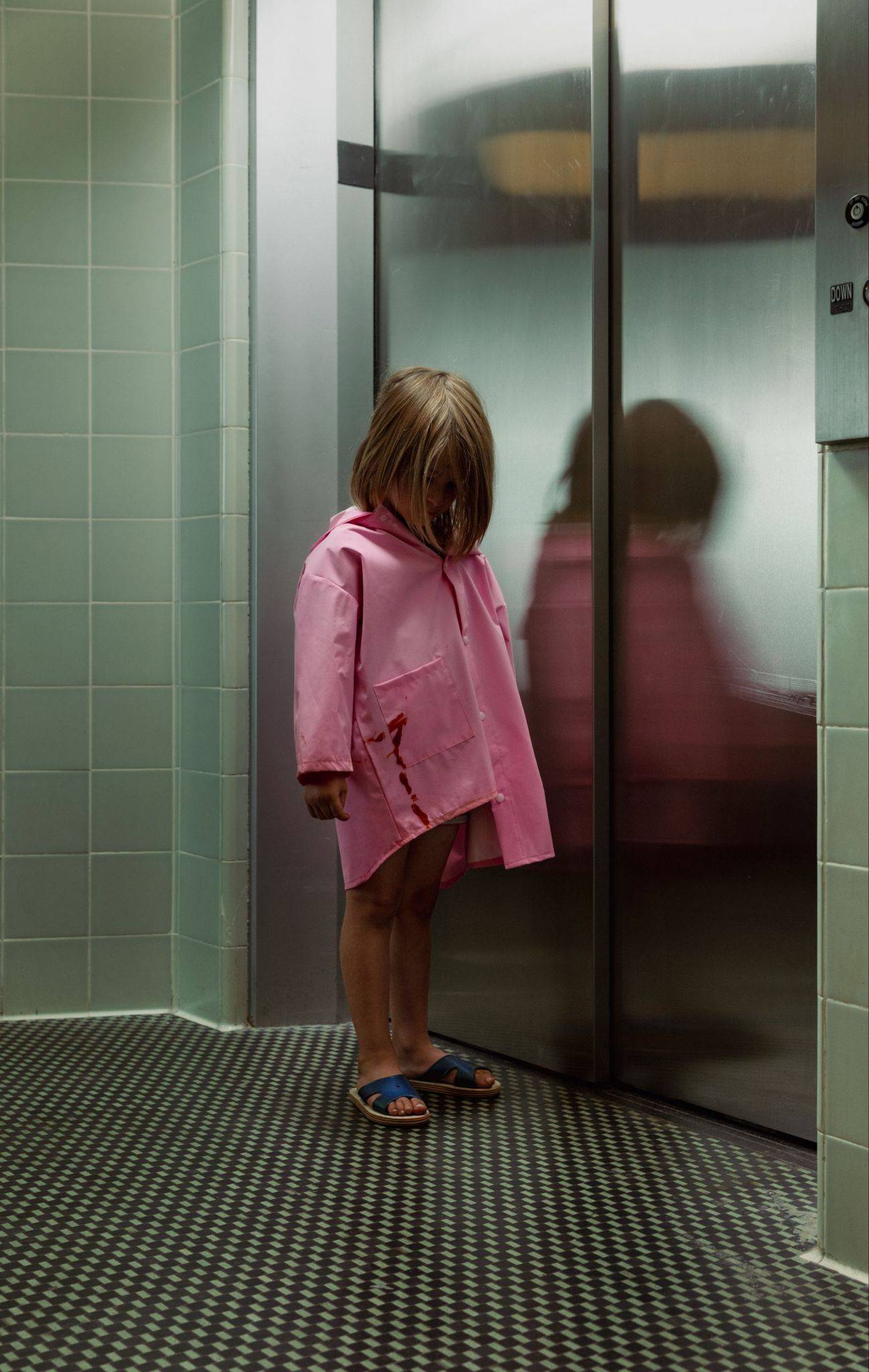 A photograph of a young girl wearing a pink jacket standing in a tiled hallway in front of an elevator.