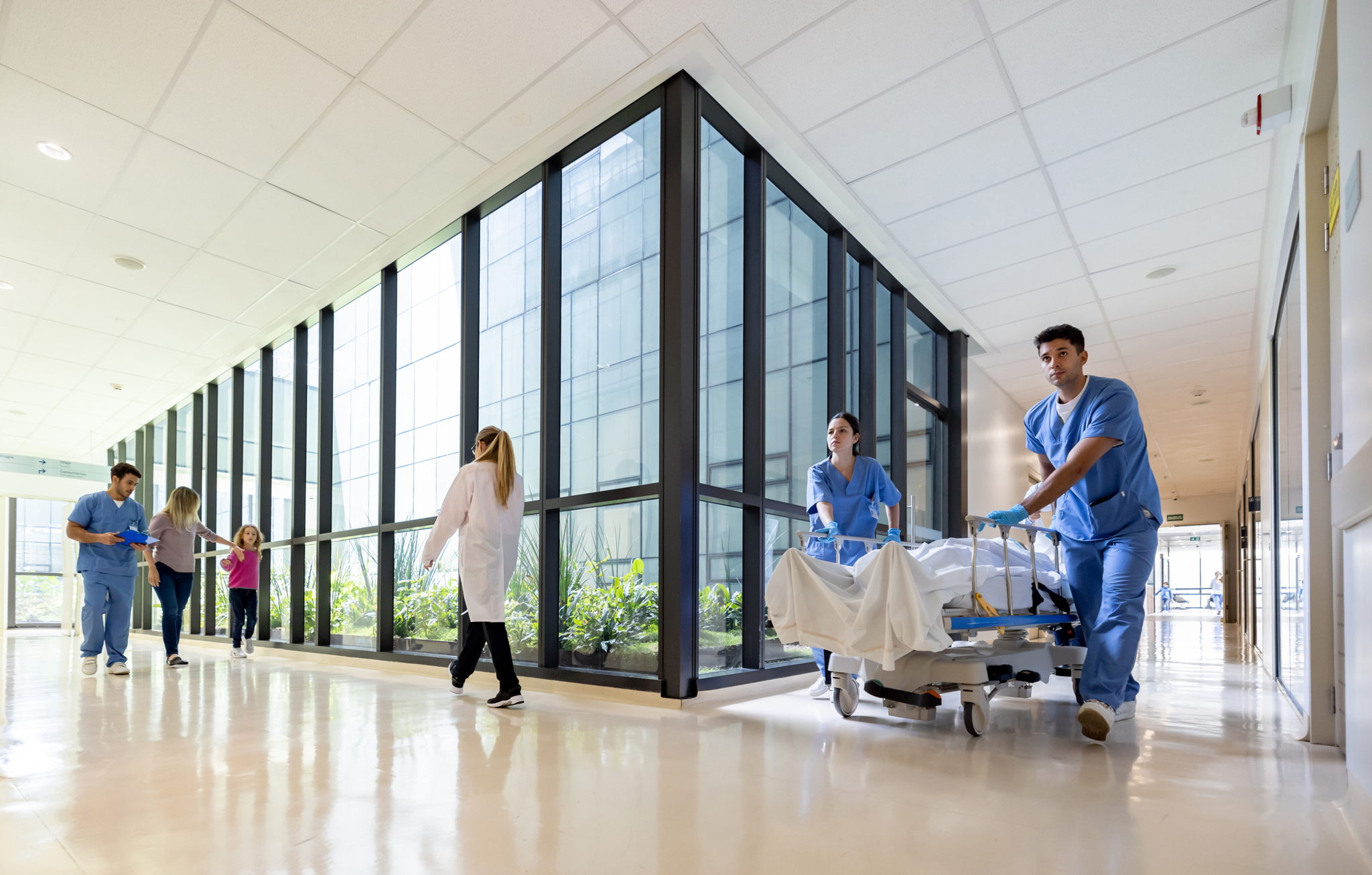 Healthcare workers in scrubs and a doctor in a white coat hurry down a bright hospital corridor, pushing a patient on a gurney. Large windows line the hallway, and two other people walk in the background. The floor is shiny and reflective.