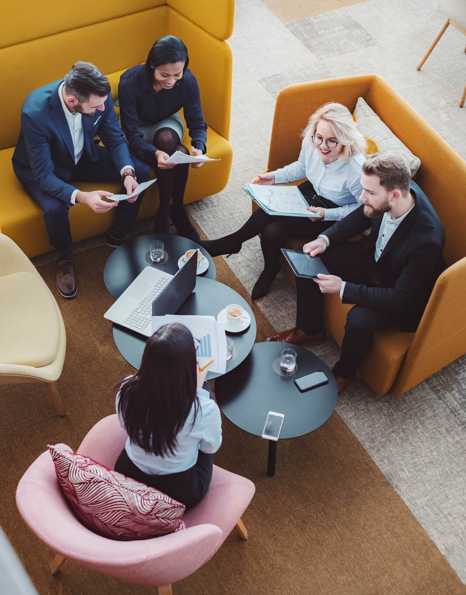 Five people are seated around two small tables in a modern office lounge, designed through our workplace services, engaging in a meeting. They are holding documents and using a laptop and tablet. The furniture includes a sofa, armchair, and coffee tables. Coffee cups are on the tables.