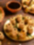 Earthen pot–shaped Indian sweets arranged on a golden tray, with honey being drizzled from a wooden dipper and scattered nuts in the background.