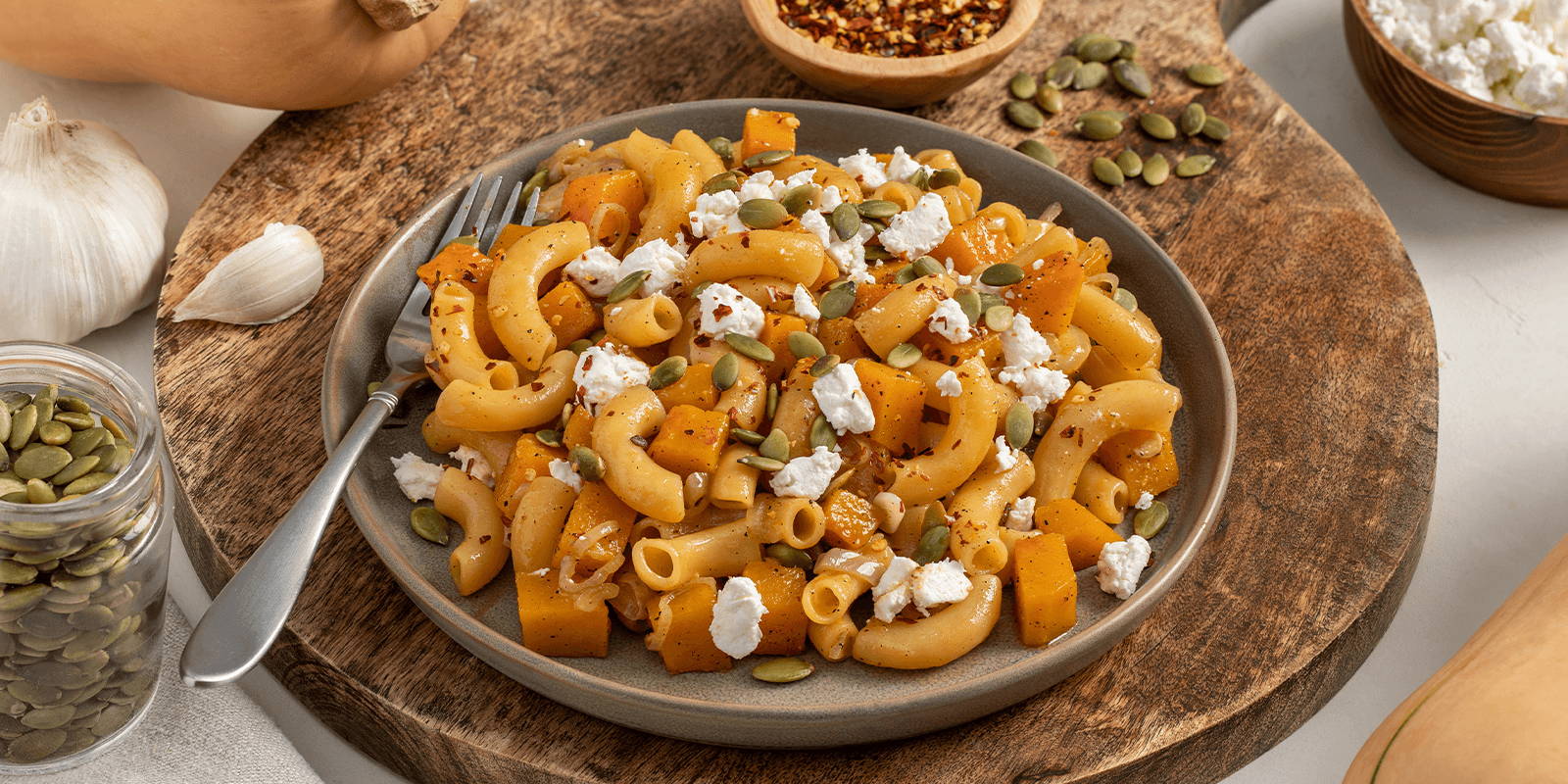A gray plateful of ZENB Elbows Pasta With Butternut Squash & Goat Cheese atop a round wooden board, styled next to its whole ingredients.
