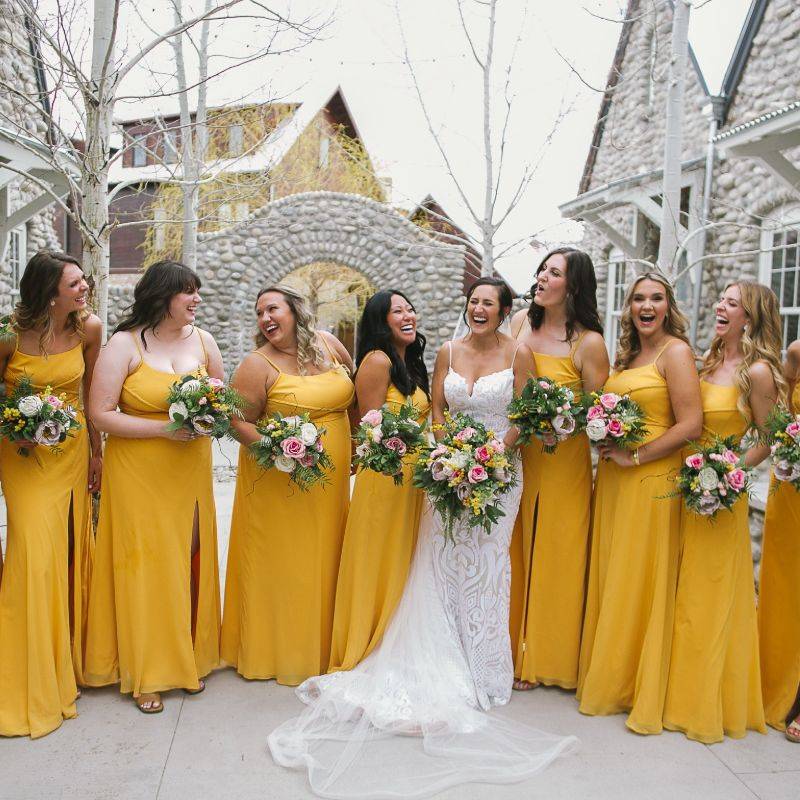 A bride wearing a white wedding dress holding a cascading bouquet made of dusty rose roses, yellow mimosa, and eucalyptus is surrounded by her bridesmaids wearing yellow dresses and holding smaller bouquets containing the same flowers