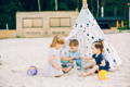 Three children enjoying summer-themed sensory play with sand, buckets, and toys beside a beach tent.
