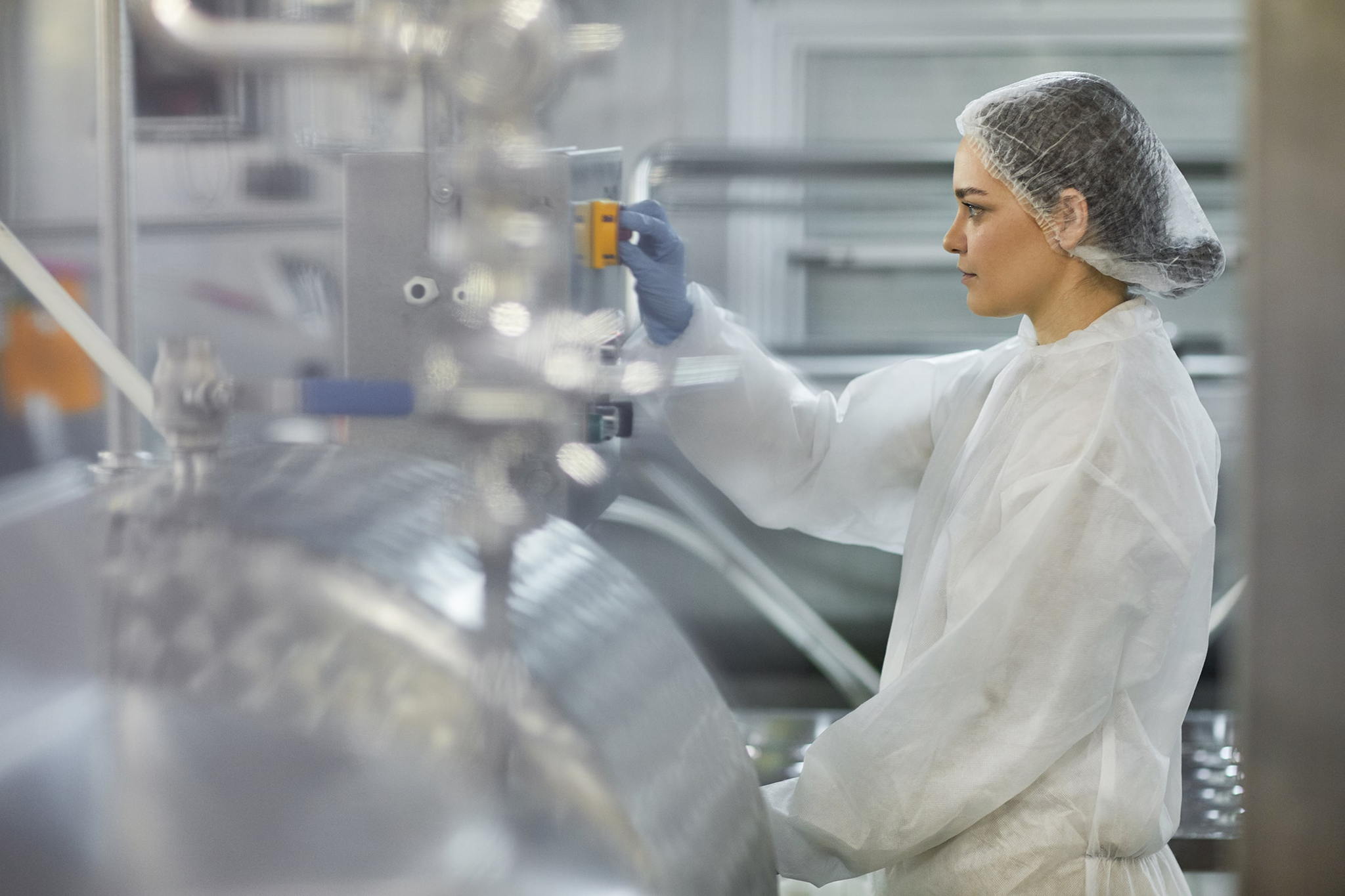 A woman in protective clothing and a hairnet operates machinery in a clean, industrial facility, likely a food or pharmaceutical processing plant. She is focused on adjusting equipment controls.