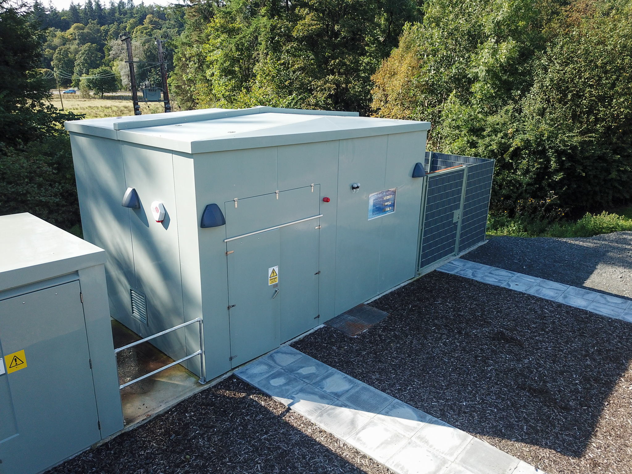 A large, light gray utility building with warning signs is situated outdoors on a gravel surface, surrounded by green trees and a wire fence. A concrete walkway runs beside the structure.