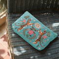 A woman in a white dress standing in speckled sunlight against an old wooden door. She is holding an Elizabeth Scarlett velvet embroidered wash bag in a turtle design, with a mid blue background.