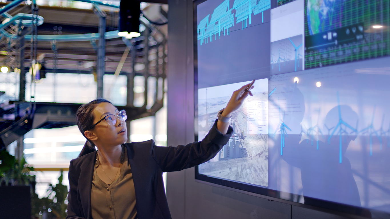 A woman in business attire points at a large digital screen displaying charts, graphs, and images in a modern office or control room setting.