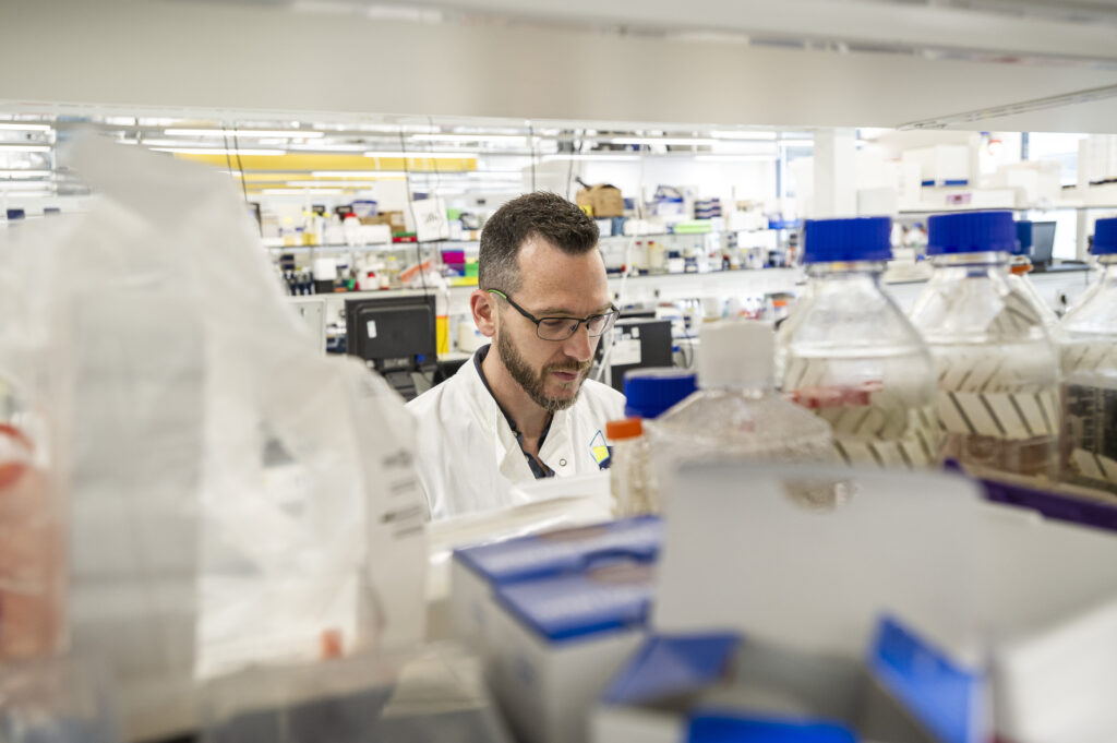 A scientist in a white lab coat works in a bright, modern laboratory, surrounded by bottles, boxes, and lab equipment on shelves and benches.