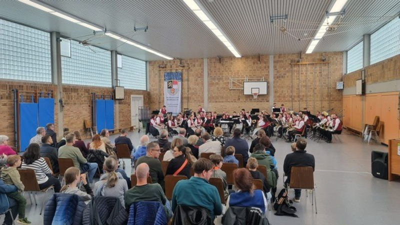 In einer Turnhalle sitzt ein Blasorchester vor Publikum und musiziert. 