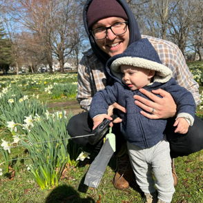 A man smiles holding a child in a daffodil field, evoking joy and warmth on a sunny, chilly spring day.