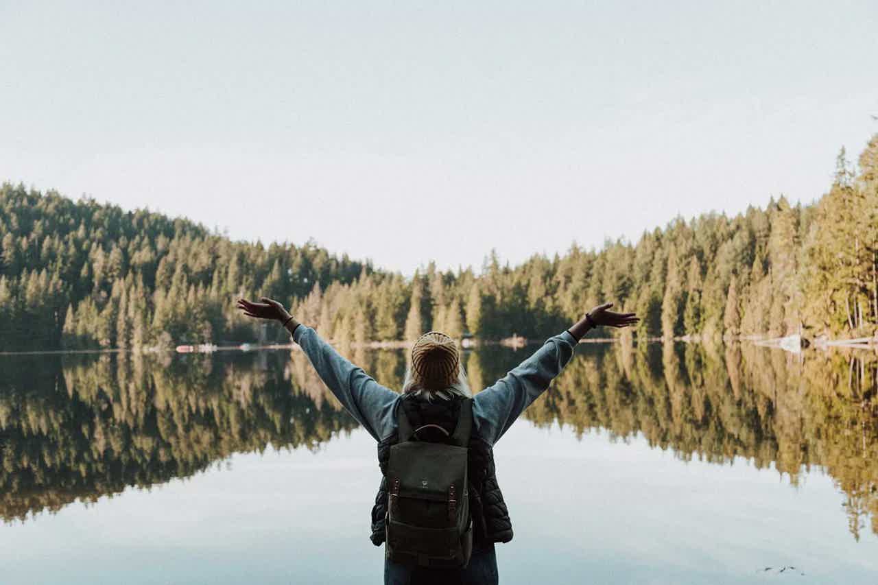 Person with backpack stands arms out by calm lake, evergreen forest reflections in misty sky.