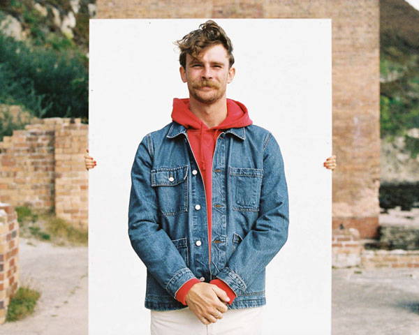 Man wearing blue denim workwear jacket with red hoodie beneath from the sustainable clothing brand Paynter based in the UK