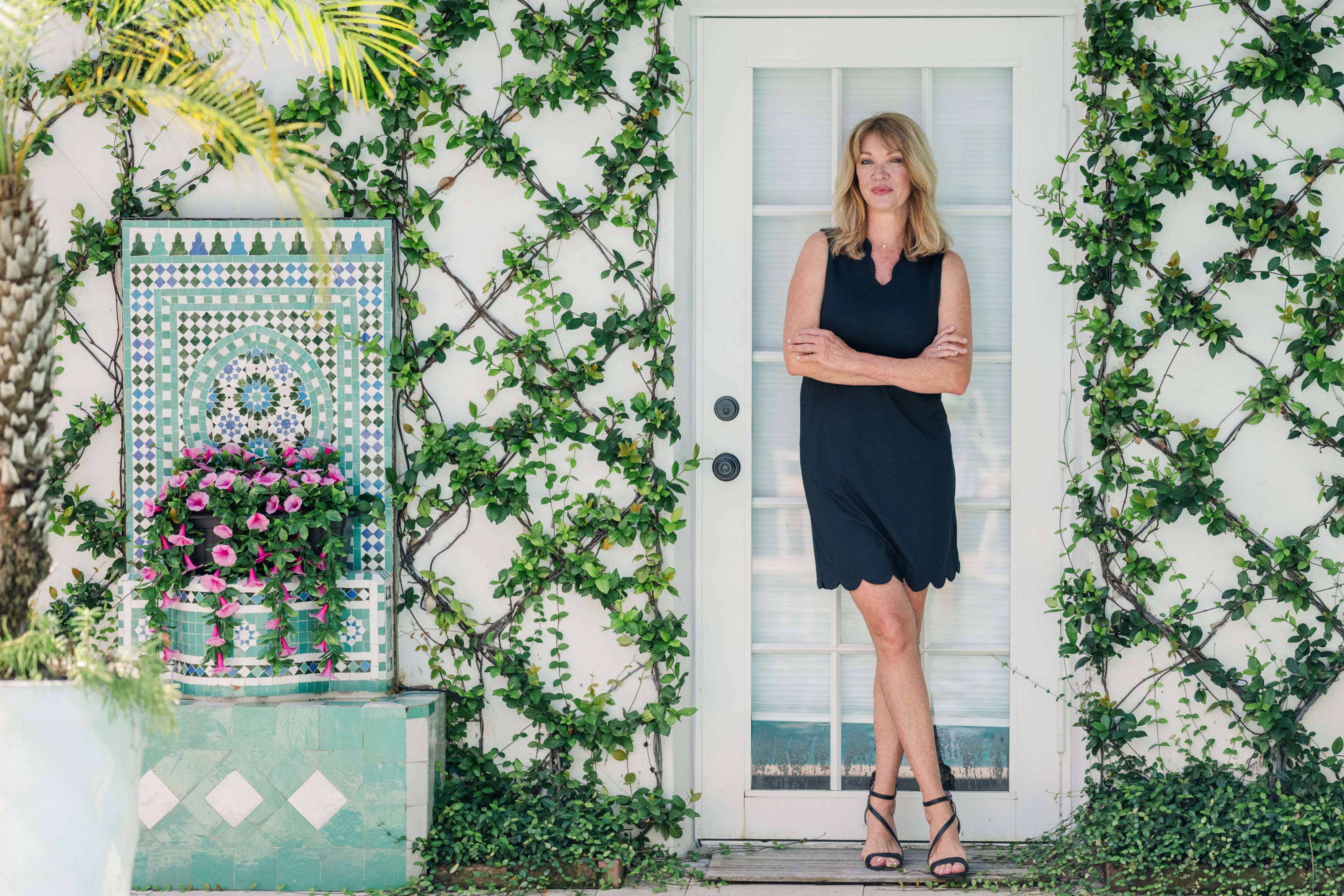 A woman in a black dress stands in front of a door in a garden with a fountain