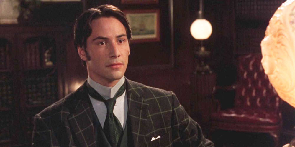 A baby faced Keanu Reeves wearing a victorian era suit in a dimly lit room.