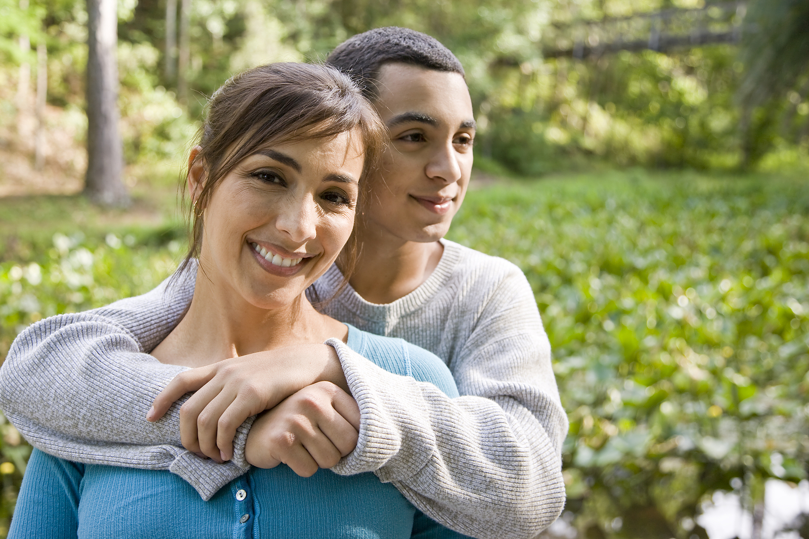 A latin mom smiles as her son has his arms around her and smiles while they are outdoors.